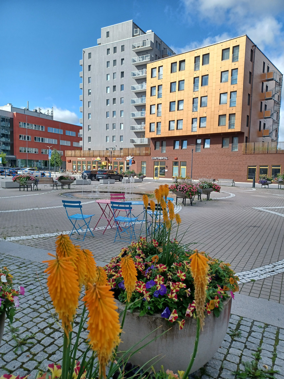 Blommor och utemöbler på Sågverkstorget i förgrunden; parkering, flervåningshus och kommunhuset i bakgrunden.