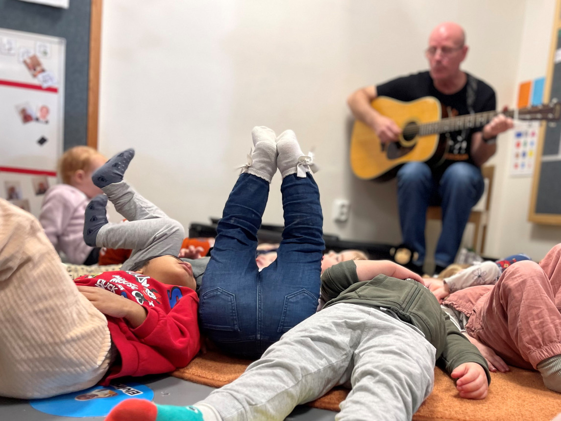 I förskolemiljö. Barn ligger på golvet, en del med ben och fötter i vädret. I bakgrunden sitter en man med gitarr.