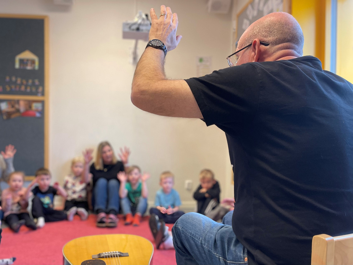Inne på en förskola. En man håller upp en hand i luften och dirigerar barn som sitter på golvet med en förskollärare. På golvet ligger en gitarr.
