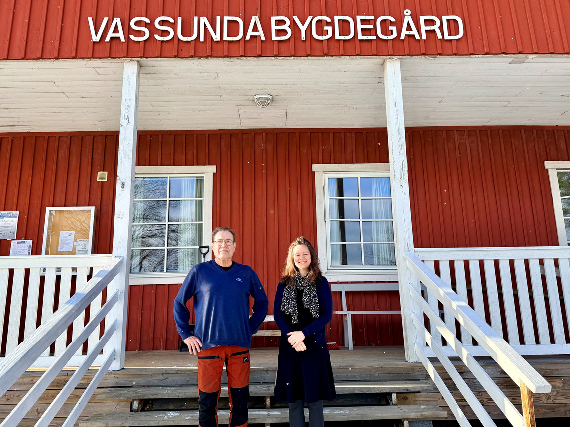 En man och en kvinna står på en trappa utanför en röd byggnad med texten Vassunda Bygdegård