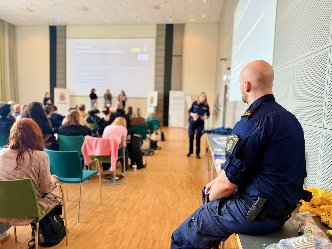 En manlig polis sitter på ett bord och tittar på tre personer som står på en scen och presenterar. I rummet finns flera människor som sitter på stolar.