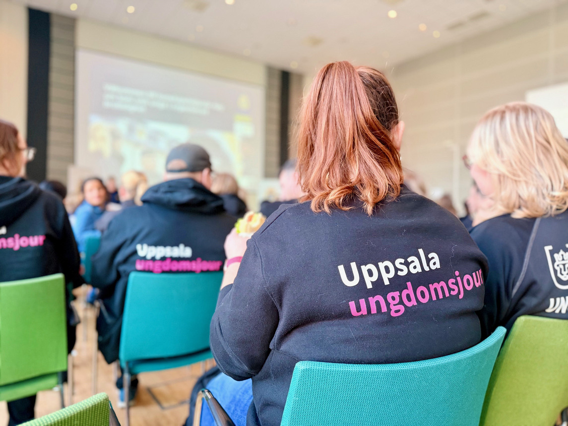 Personer som sitter på stolar i en sal och ser en presentation - på deras ryggar står det Uppsala ungdomsjour.