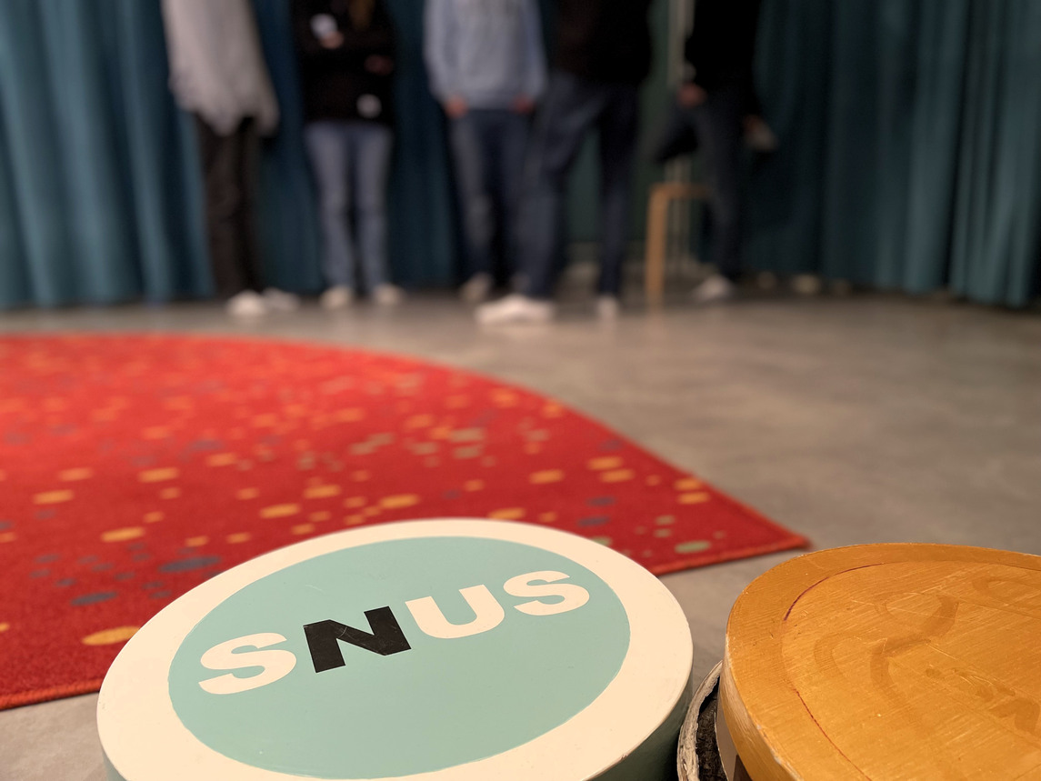 En stor mintgrön dosa som det står snus på - i bakgrunden fem ungdomar i jeans.