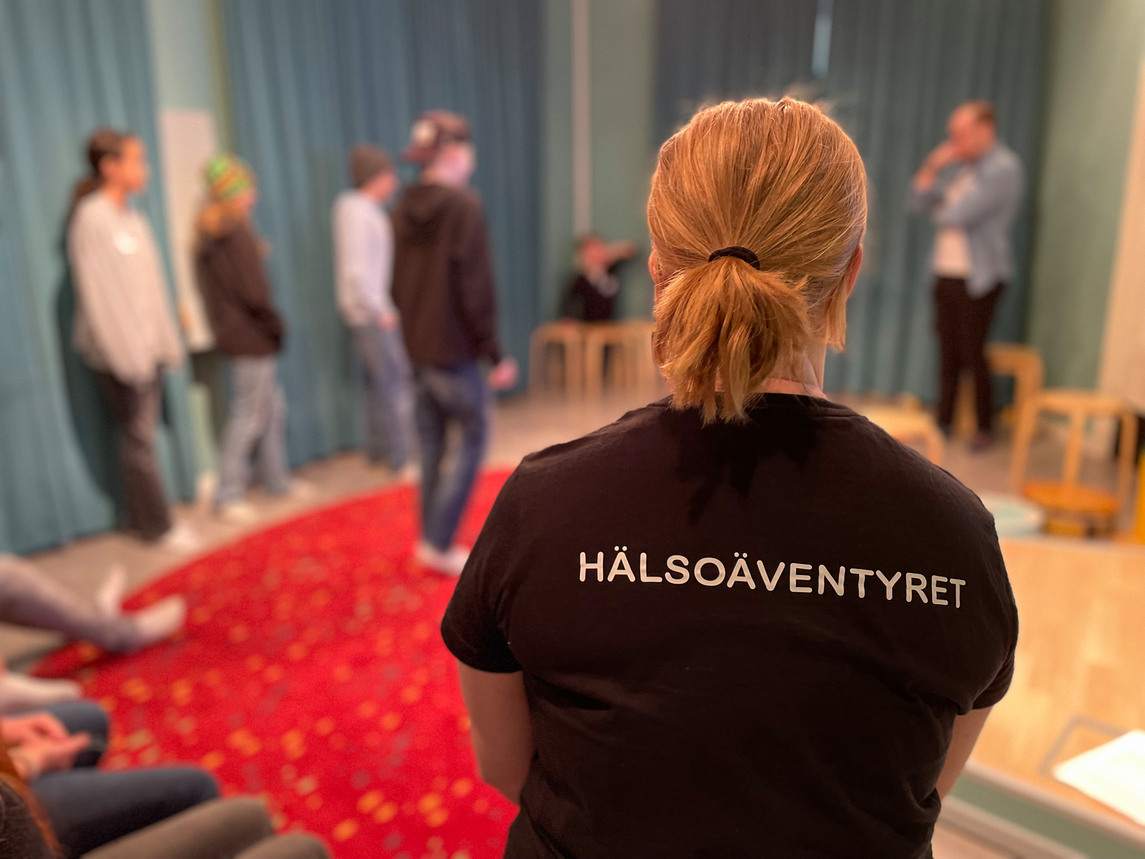 En kvinna med svart t-shirt med texten Hälsoäventyret i förgrunden - i bakgrunden ungdomar som gör en teaterscen