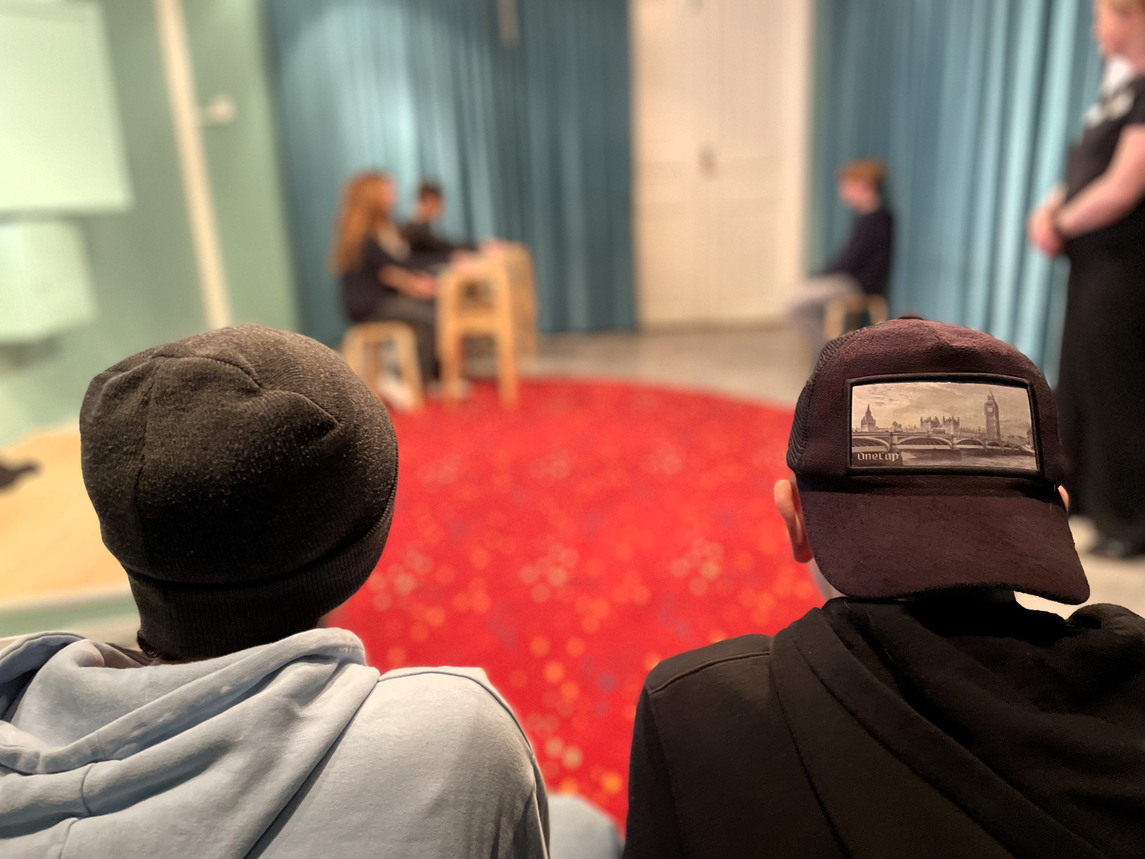 Två ungdomar i keps och mössa som tittar på kamrater som spelar teater.