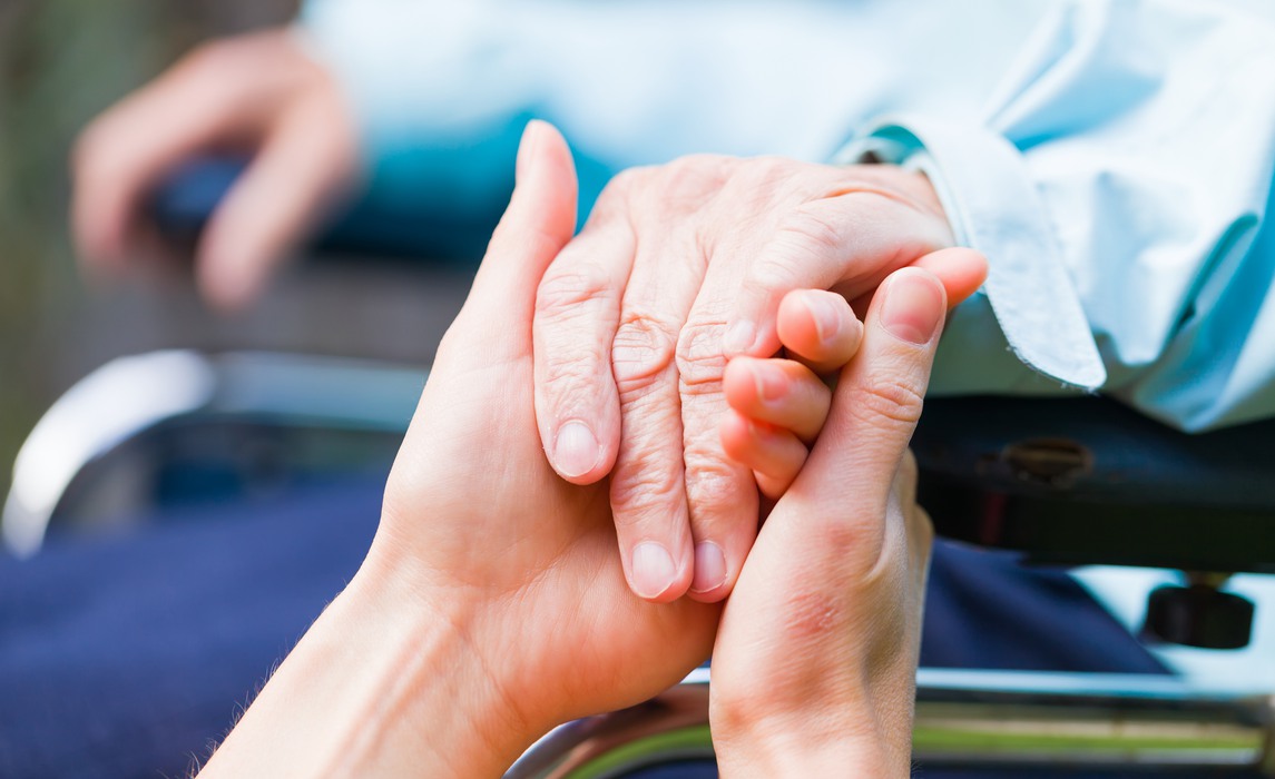 Yngre vårdgivare håller hand på äldre person i rullstol