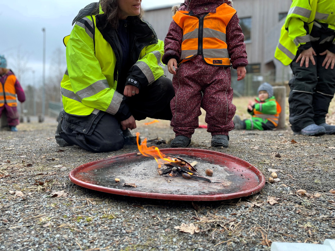 Ett barn i lila overall och orange väst som står framme vid ett eldfat. Bredvid sitter en förskollärare på huk.